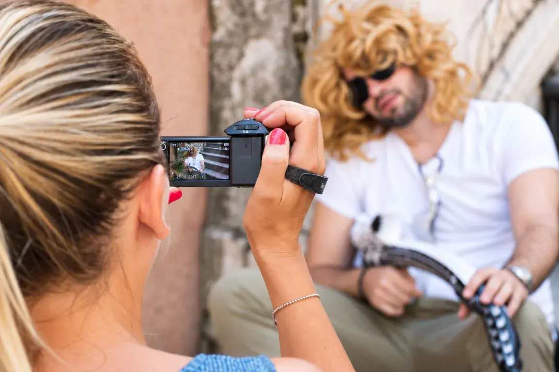 A team member filming a colleague in a blonde wig and sunglasses with a camcorder on a Sarajevo street