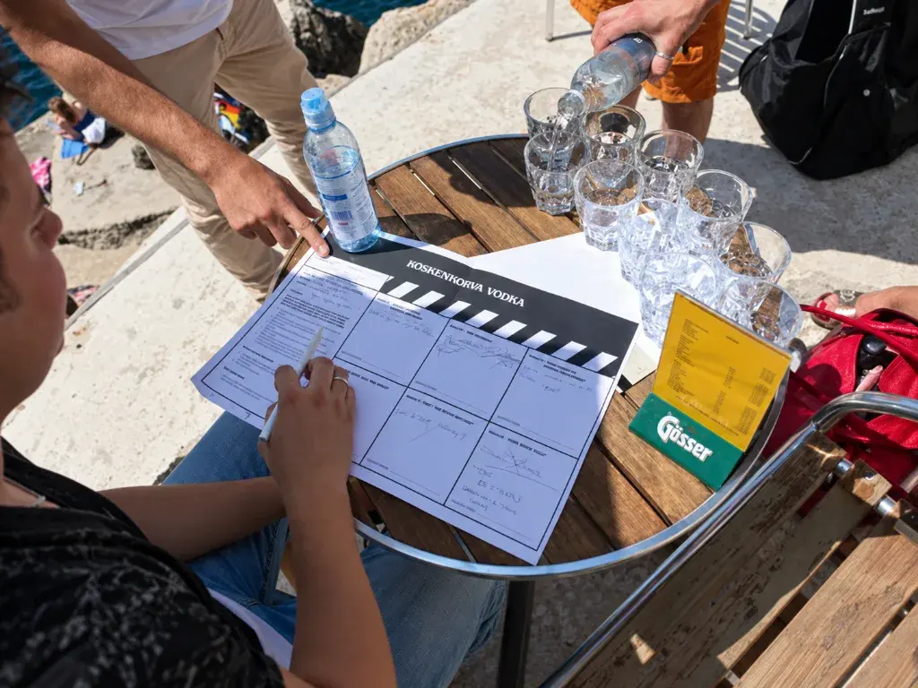 A team member filling in a clapperboard-style shot list at an outdoor table while water is poured into glasses