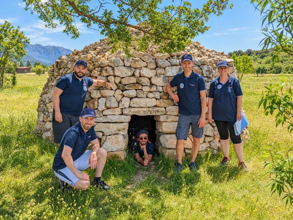 Colleagues in matching polo shirts posing around a stone hut, one peeking out from inside
